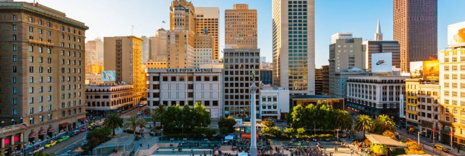 Aerial view of a busy city plaza with a tall central monument, palm trees, and surrounding high-rise buildings filled with pedestrians.